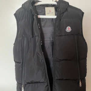 Svart dunväst från Moncler med huva - Svart dunväst från Moncler med broderad logga på bröstet och stor vit Moncler-text på insidan av huvan. Västen har dragkedjor framtill och på fickorna samt en quiltad design. Perfekt för lager-på-lager och streetstyle. KOM GÄRNA MED EGNA PRISFÖRSLAG!