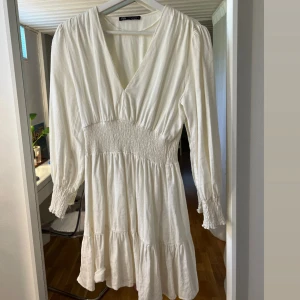 Vit kortklänning från Zara - Superfin vit kortklänning från Zara med v-ringning, smockad midja och långa ärmar med smock vid ärmslut. Klänningen har volangdetaljer och är tillverkad i ett mjukt bomullstyg. Perfekt för sommarens alla tillfällen!💓