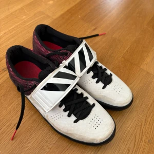 Adidas Adizero träningsskor vit/svart KULSTÖTNING - Säljer ett par Adidas Adizero träningsskor i vitt och svart med svarta snören och bred kardborre över foten. Ovandelen har mesh i rött och svart, och sulan är helsvart. Friidrott Kulstötning 