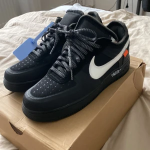 Nike Air Force 1 Off-White svart/vit - Nike Air Force 1 Off-White i svart mesh och syntet med vit swoosh, svarta och vita snören och klassiska Off-White detaljer som blå tag och orange patch. Platt sula och rund tå. Snygg streetwear-vibe och ikonisk design.