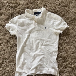Vit pikétröja Polo Ralph Lauren - Klassisk vit pikétröja från Polo Ralph Lauren i slim fit-modell. Tröjan har korta ärmar, krage med knappar och den ikoniska broderade loggan på bröstet. Perfekt för en clean och stilren look. Tillverkad i mjuk bomull.
