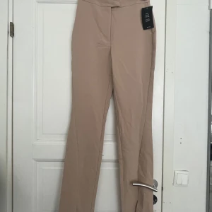 Beiga kostymbyxor från NA-KD, strl 34 - Snygga beiga kostymbyxor från NA-KD med hög midja och raka ben. Byxorna har en stilren look och är gjorda i ett mjukt material som känns skönt mot huden. Perfekta att styla med en crop top eller oversized skjorta.