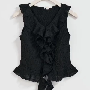 Super snygg svart volangblus - Supersnygg svart blus med v-ringning och volangdetaljer framtill. Ärmlös modell i lätt, krinklat tyg som ger en cool struktur. Toppen är storlek 158 164 men passra som xs. Passar till både jeans och kjol för en trendig look. Liten bitt av tyget är borta på en av ändarna (syns ej när man har på sig den) se sista bilden 💞