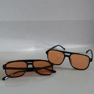 Svarta pilotbågar med orange glas - Två par stilrena solglasögon med svarta pilotbågar och rektangulära orangefärgade glas. Bågarna är i plast och har en klassisk dubbelbrygga. Perfekta för dig som vill sticka ut med en retrovibe och cool färg på linserna. Kan köpas separat för 150
