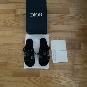 Svarta sandaler från Dior  - Snygga svarta sandaler från Dior med tydligt logomönster i vitt och svart. Skorna har breda remmar i textil och en platt sula. Perfekta för dig som vill ha en lyxig och trendig look med streetkänsla. Kommer med originalkartong och dustbag.