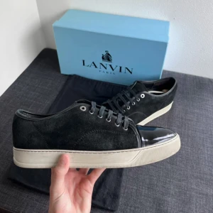 Svarta sneakers från Lanvin med lackad tå - Stilrena svarta sneakers från Lanvin med ovandel i mocka och en glansig, lackad tå. Skorna har vita platta sulor och svarta snören. Perfekt för dig som gillar en clean look med exklusiva detaljer.