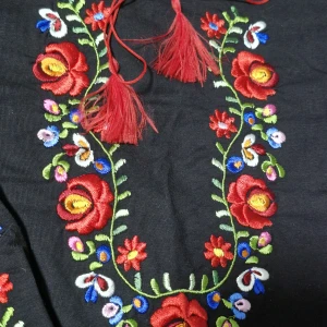 Svart blus med broderade blommor - Svart långärmad blus med färgglada broderade blommor i rött, blått, grönt och vitt längs halsringningen och ärmarna. Dekorativ snörning med röda tofsar vid halsen och röda sicksack-detaljer vid ärmslut. Tillverkad i mjuk bomull.