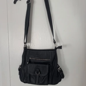 Svart axelväska med detaljer - Svart axelväska i skinnimitation med justerbar rem, silvriga spännen och dragkedjor. Väskan har en framficka med spänne och lock, samt vita kontrastsömmar som ger en cool look. Perfekt storlek för att få plats med allt du behöver till vardagen.