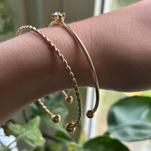 Två guldiga armband med sten - Säljer två snygga armband/bangles i guldig metall. Det ena är tvinnat och har små kulor i ändarna, det andra är slätt med en klar kristall på ovansidan. Perfekta att bära tillsammans för en stilren look.