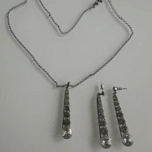 Glittrigt smyckeset med halsband & örhängen - Snyggt smyckeset med halsband och matchande örhängen i silverfärg. Både halsbandet och örhängena har avlånga hängen med flera glittrande stenar och en större kristall längst ner