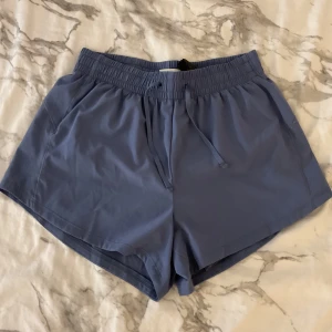 Blå träningsshorts med snörning - Snygga blå träningsshorts med elastisk midja och snörning framtill. De har en loose passform och är tillverkade i ett lätt syntetmaterial som andas, perfekta för aktiva dagar. Shortsen är midwaist och har en enkel, stilren design utan mönster.
