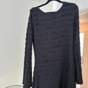 Svart kortklänning med öppen rygg - Snygg svart kortklänning med lång ärm och cool struktur i tyget. Klänningen har en öppen rygg som ger en extra edge och är tillverkad i ett stretchigt material. Perfekt för dig som vill sticka ut med en unik look.