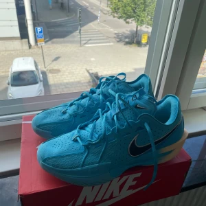 Blue Fury-Black-Mtlc Silver - Snygga blå Nike basketskor med svart swoosh och vita detaljer. Ovandelen är i ventilerande mesh och syntet, med perforeringar för extra andningsförmåga. Skorna har snörning och en platt sula i beige och blått, perfekt för dig som gillar sportig stil och även spelar basket.