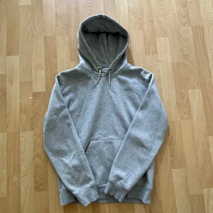Grå hoodie från Carhartt - Säljer en stilren grå hoodie från Carhartt med broderad logga i guld på ärmen. Tröjan har huva med snörning, känguruficka och är tillverkad i mjuk bomullsmix. Perfekt för chill dagar eller när du vill ha en clean streetwear-look.