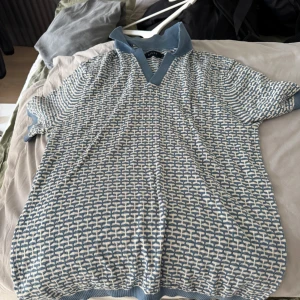 Blåvit stickad pikétröja från Riley - Snygg blåvit stickad pikétröja från Riley med korta ärmar och mönstrad design. Tröjan har en klassisk krage och v-ringning i blått, samt ribbade muddar vid ärmslut och nederkant. Perfekt för dig som gillar retrovibbar och unik stil. Ny pris 699kr