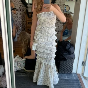 Vit blommig långklänning med volanger - Superfin axelbandslös långklänning i vitt med blommigt mönster🥰 jag är 168 och den passar perfekt på mig i längden! Har endast använt den en gång 