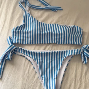 Randig blåvit bikini från SHEIN - Superfin bikini från SHEIN i blått och vitt med vertikala ränder. Toppen är asymmetrisk med en axel och knytband, medan underdelen har knyt i sidorna. Tillverkad i stretchig polyesterblandning som sitter snyggt och bekvämt. Perfekt för stranden eller poolen!