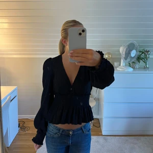 Mörkblå blus med volang från NA-KD - Supersnygg mörkblå blus från NA-KD med v-ringning, smockad midja och volangkant nertill. Långa puffärmar med smock vid ärmslut och lätt struktur i tyget. Perfekt att styla med jeans eller kjol för en trendig look. Hör av er vid frågor 💕💕