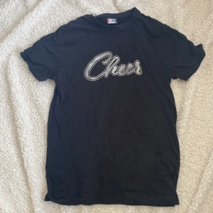 Svart t-shirt med glittertext från Clique - Svart t-shirt från Clique med coolt tryck där det står 'Cheer' i glittriga bokstäver och små silvriga nitar runtom. Klassisk passform och rund halsringning. Perfekt för dig som gillar statement-plagg och vill sticka ut lite extra. använd typ fyra gånger.