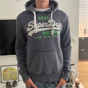 Mörkgrå Superdry hoodie med tryck - Säljer en mörkgrå hoodie från Superdry med vit och grön logga framtill. Tröjan har huva med vita snören, känguruficka och ribbade muddar. Perfekt för dig som gillar en avslappnad och sportig stil.
