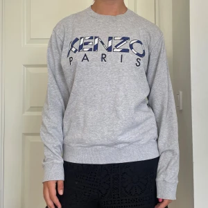 Grå Kenzo Paris sweatshirt - Säljer en grå sweatshirt från Kenzo Paris med stort broderat logotyptryck i blått och vitt framtill. Tröjan har rund hals, ribbade muddar och är långärmad. Perfekt för dig som gillar stilrena och trendiga plagg med streetkänsla. Skriv om du undrar något, kolla min profil innan du trycker på köp nu💗💗