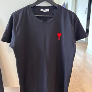 Svart t-shirt från Ami med rött hjärta - Svart t-shirt från Ami med klassisk passform och rund hals. På bröstet finns ett broderat rött hjärta med bokstaven A. Tillverkad i mjuk bomull och har korta ärmar. Perfekt för en clean och stilren look. Passar medium.