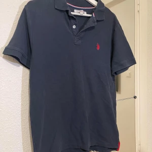 Mörkblå pikétröja från US Polo Assn - Klassisk mörkblå pikétröja från US Polo Assn med röd broderad logga på bröstet. Tröjan har korta ärmar, krage och knäppning med två knappar. Perfekt för en stilren och avslappnad look.