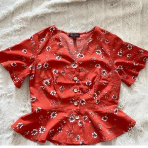 Röd blommig blus från QED London - Superfin röd blus från QED London med vita och lila blommor, v-ringning och knappar framtill. Blusen har korta ärmar och volang nertill som ger en söt och trendig look. Perfekt för dig som gillar färg och mönster!