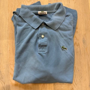 Ljusblå pikétröja från Lacoste - Snygg ljusblå pikétröja från Lacoste med klassisk krage och två knappar framtill. Ikoniska broderade krokodilloggan på bröstet. Tröjan är i bomull och har en tidlös, clean look som passar perfekt till jeans eller shorts.