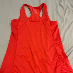 Röd linnetopp från Newbody - Snygg röd linnetopp från Newbody med brottarrygg. Perfekt för träning eller när du vill ha en sportig look. Tillverkad i ett lätt och stretchigt syntetmaterial som andas. Enkel design utan mönster, sitter skönt och följer kroppen.