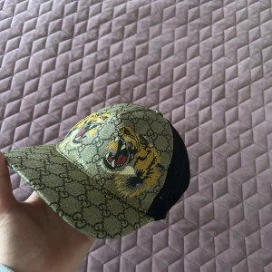 Gucci keps med tigertryck och GG-mönster - Snygg Gucci keps i beige med klassiskt GG-mönster och två stora tigerhuvuden framtill. Baksidan är i svart mesh för extra ventilation och justerbar passform. Ikoniska gröna och röda Gucci-ränder vid spännet. Perfekt statement-piece för dig som vill sticka ut.