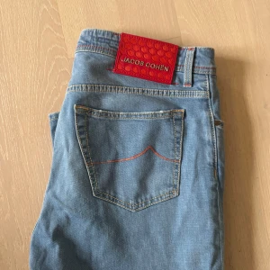 Jacob Cohen ljusblå jeans Bard 35 - Snygga ljusblå jeans från Jacob Cohen, modell Bard. Jeansen har röda kontrastsömmar, klassisk femficksdesign och en röd patch med logga bak. Tillverkade i mjukt bomullsjeans-tyg med knappgylf och unika detaljer på fickorna. Notera att jeansen är snäppet kortare än vanliga cohens, be om mått. Snabb affär prioriteras. 
