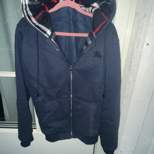 Blå Burberry zip up - Säljer en blå hoodiejacka från Burberry i storlek L. Jackan har dragkedja, snörning i huvan och klassiskt rutigt foder i huvan och insidan. Tillverkad i 100% bomull, med två stora fickor framtill och diskret Burberry-logga på bröstet. storlek L passar även dig med M!☺️