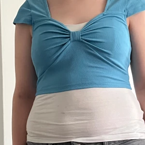 Blå croppad ribbad topp med twist - Säljer en blå croppad topp med korta ärmar och snygg twistdetalj framtill. Toppen är ribbad och har en tight passform som framhäver figuren. Perfekt för dig som gillar trendiga och bekväma plagg med lite edge. Det är dock lite tråd innan för tröjan som sticker fram lite. Men syns inte,finns bild här upptill ⬆️vit linne hör ej till!