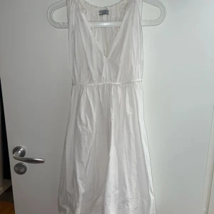Vit omlottklänning från Hunkydory - Supersöt vit omlottklänning från Hunkydory i 100% bomull. Klänningen har v-ringning, knytband i midjan och är ärmlös. Perfekt för varma dagar när du vill känna dig både chill och snygg.