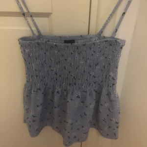 Blommig blå linnetopp Gina Tricot XS - Supersöt ljusblå linnetopp från Gina Tricot med smock upptill och tunna axelband. Toppen har ett blommigt mönster i vitt och mörkblått samt en lätt volang nedtill. Perfekt för varma dagar och ger en fräsch sommarkänsla.