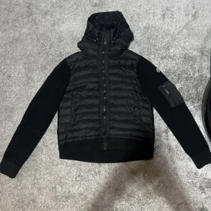 Svart Moncler pufferjacka med huva - Svart pufferjacka från Moncler med stickade ärmar och vadderad kropp. Jackan har huva, dragkedja framtill och en ficka med dragkedja på ärmen. Klassisk Moncler-logga på vänster ärm. Perfekt för kalla dagar och riktigt snygg streetstil.