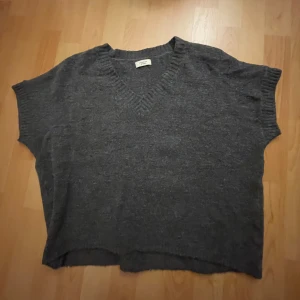 Mörkgrå stickad tröja Jacqueline de Yong - Mysig mörkgrå stickad tröja från Jacqueline de Yong med v-ringad hals och korta ärmar. Tröjan har en loose passform och ribbade detaljer vid halsringning och ärmslut. Perfekt för lager-på-lager och chill vibes.