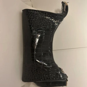 Svart axelväska i croco-imitation - Snygg svart axelväska med croco-mönstrad yta och stilren form. Väskan har dragkedja upptill och silvriga metalldetaljer. Perfekt storlek för det viktigaste och ger en cool vibe till din outfit.