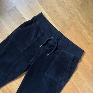 Svarta velourbyxor Juicy Couture  - Svarta mjukisbyxor från Juicy Couture i mjuk velour med dragsko i midjan och stora fickor fram. Raka ben och bakfickor med knappdetalj. Perfekta för en chill och trendig look.