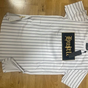 Vit randig t-shirt Supply & Demand - Vit t-shirt med svarta tunna ränder från Supply & Demand. Stor broderad patch med texten 'Hustle' i guld på svart bakgrund framtill. Kortärmad modell med rund hals och guldfärgad dragkedja i sidan. Skön streetstil med snygga detaljer.