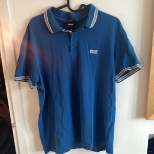 Blå pikétröja från Hugo Boss - Snygg blå pikétröja från Hugo Boss med vit logga på bröstet. Tröjan har en klassisk krage med vita ränder och knappar framtill. Perfekt för en stilren look.                                          Storlek: Xl men passar M