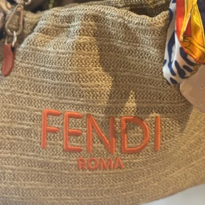 Beige axelväska från Fendi med orange detaljer - Säljer en beige axelväska från Fendi med broderad logga i orange och bred axelrem med orange kant. Väskan är flätad i ett naturmaterial och har snygga läderdetaljer samt en färgglad scarf knuten i ena sidan. Perfekt för dig som vill sticka ut med en lyxig och trendig accessoar.