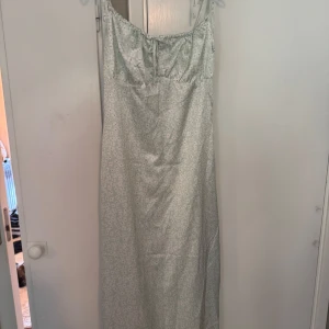 Ljusblommig långklänning med knyt - milkmaid modell - Superfin långklänning i ljusgrönt siden tyg med små vita blommor och rosa prickar. Klänningen har smala axelband och söt knytning framtill vid bysten so ger milkmaid modell. Perfekt flowy modell för varma dagar och somriga vibes.