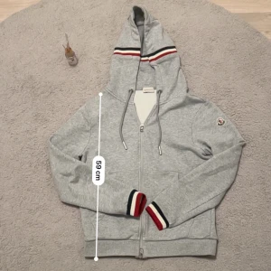 Grå Moncler hoodie med dragkedja - Snygg grå hoodie från Moncler med hel dragkedja, huva och snörning. Ribbade muddar med röd, vit och marinblå detalj samt matchande rand på huvan. Klassisk Moncler-logga på ärmen. .Jag är 181 och väger 65 kg, den passar mig perfekt.Den är i storlek M men passar S också, se även mått på bild 1 och 2. Den har ett litet rev vid bild 4 men annars är det bra kvalitet.Tröjan passar perfekt för chill dagar men även hösten 🍂 och vintern ❄️. 💬 Skriv gärna om du har frågor! 📦 Skickas inom 24 timmar! 
