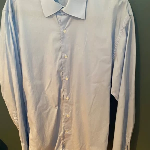 Ljusblå skjorta från Trussardi - Snygg ljusblå skjorta från Trussardi Collection med klassisk krage och knäppning framtill. Skjortan är långärmad och tillverkad i mjuk bomull, perfekt för en clean och stilren look. Passar dig som gillar enkel och tidlös stil.