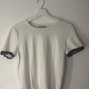 Vit t-shirt med spetsdetalj från Zara - Vit t-shirt från Zara i mjuk bomull med korta ärmar. Ärmsluten har snygga svarta spetsdetaljer som ger en cool kontrast och unik touch. Perfekt för dig som gillar stilrena plagg med en twist.