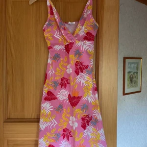 Rosa blommig klänning - Snygg klänning köpt i Spanien i rosa med tropiskt blommigt mönster i rött, gult, vitt och blått. Klänningen har smala axelband och v-ringning framtill. Perfekt för sommarens varma dagar och ger en färgglad, chill vibe. Ingen storlek står men skulle uppskatta att den är i S