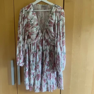 Plisserad blommig klänning H&M M - Superfin plisserad kortklänning från H&M med blommigt mönster i rosa, grönt och vitt. Djup v-ringning, långa ärmar och flowig passform.