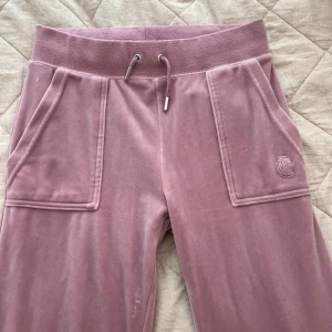 Rosa velourbyxor Juicy Couture XS - Supermjuka rosa velourbyxor från Juicy Couture med bred resår och snörning i midjan. Stora fickor fram och bak, varav bakfickorna har knappar. Klassisk broderad JC-logga på ena fickan. Perfekta för en chill och trendig look. Aldrig använda